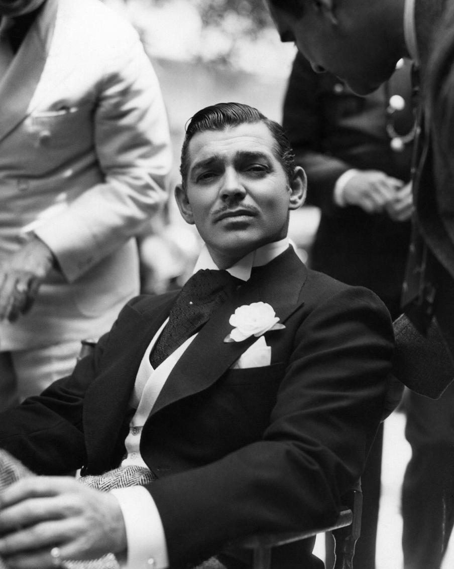 На снимке Кларк Гейбл отдыхает между сценами на съёмочной площадке фильма "Любовь в бегах". На актёре шикарный чёрный пиджак с белой розой в петлице. Жилет на Гейбле тоже белого цвета, а вот брюки, напротив, контрастные – серые, твидовые, с узором "ёлочка".