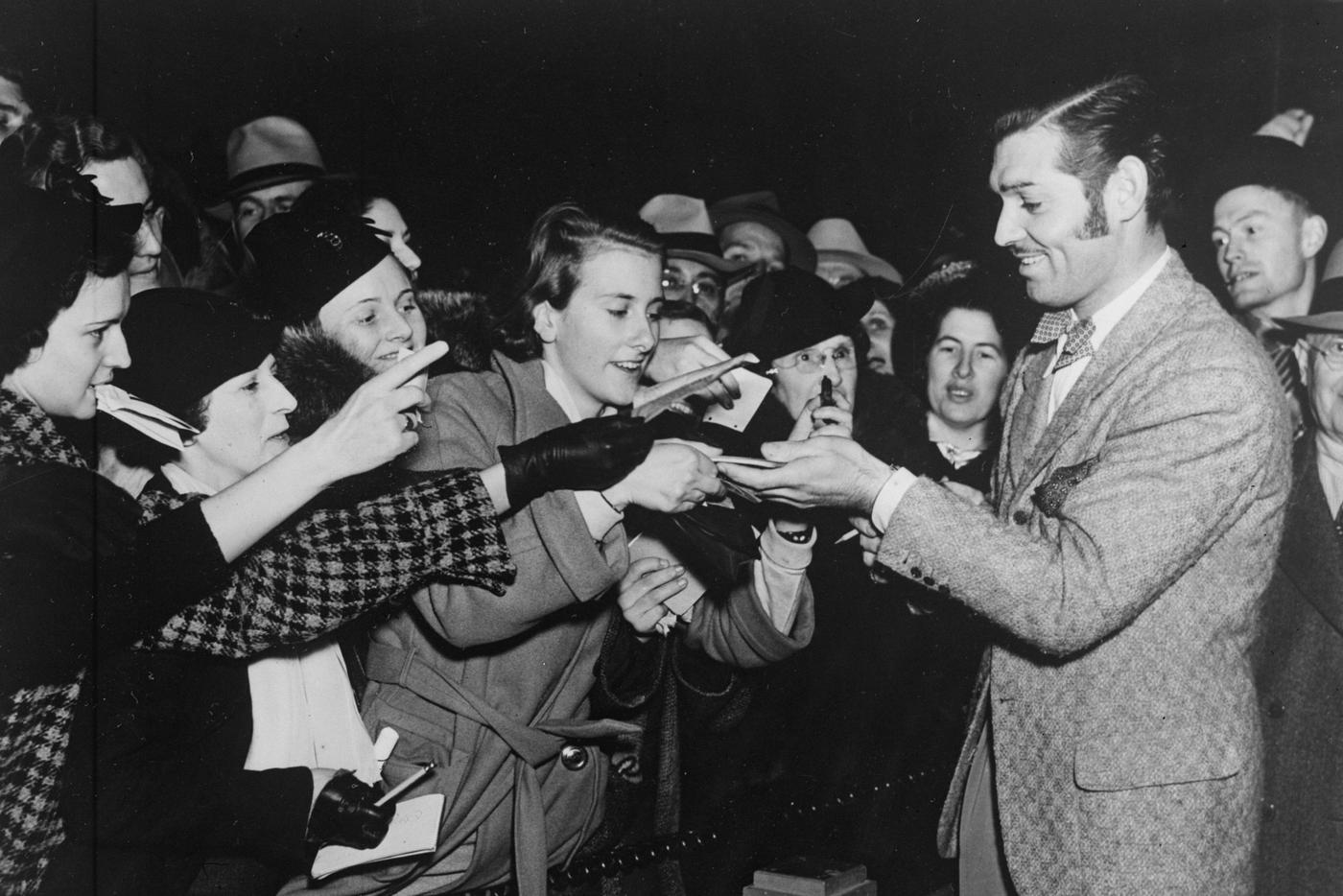 На снимке Кларк Гейбл раздаёт автографы фанатам. На нём контрастный пиджак в клетку и галстук-бабочка.