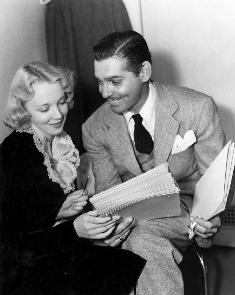 На снимке Кларк Гейбл беседует с Вирджинией Брюс, знаменитой кинозвездой Paramount, танцующей чечетку. На Гейбле элегантный светло-серый костюм-тройка в полоску.