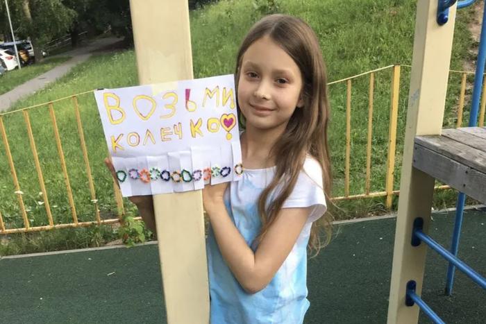 Девочка украшет район добрыми колечками из бисера. Она развешивает их во дворе с подписью "Возьми колечко"